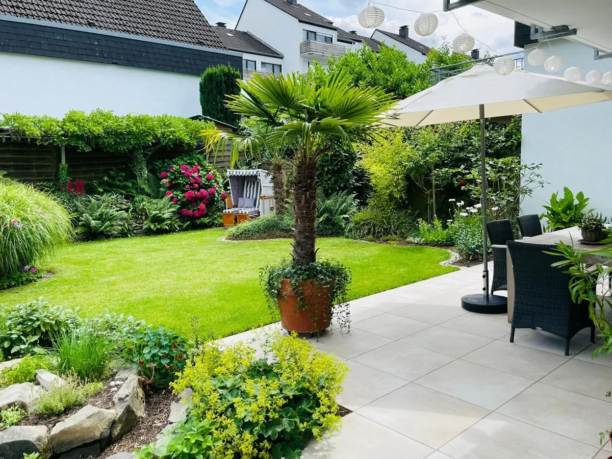 Großzügige Feinsteinzeug-Terrasse mit Palmen, Beeten und grüner Rasenfläche